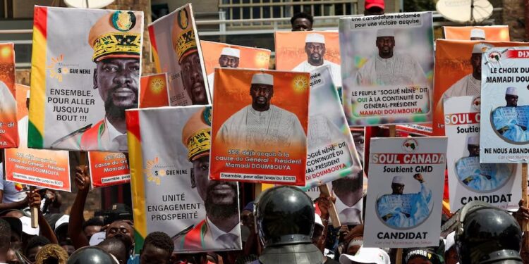 Police officers stand guard in front of supporters of Guinean leader Mamadi Doumbouya who hold campaign signs, as they wait for him to submit his candidacy at the Supreme Court ahead of the presidential election scheduled for December 28, in Conakry, Guinea November 3, 2025.